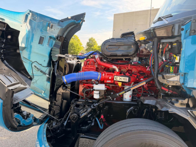 Truck engine bay of a 2024 Kenworth T680 showing a red EXIS engine with blue hoses and exposed components between the cab sections.