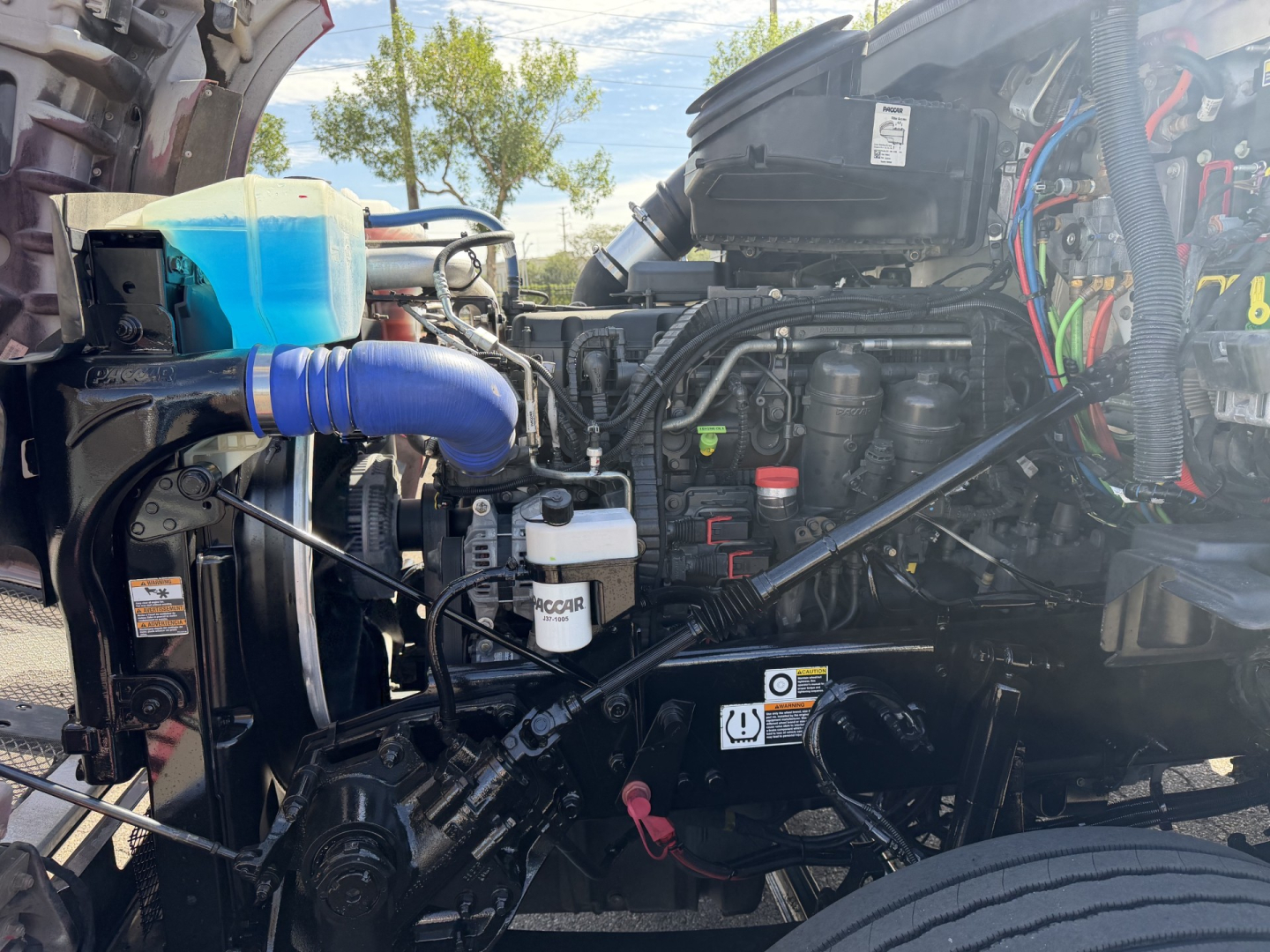 Kenworth T880 engine bay displaying coolant reservoir, hoses, filters, and engine components with visible wiring harnesses and a white ROCAIR or similar label on a filter.
