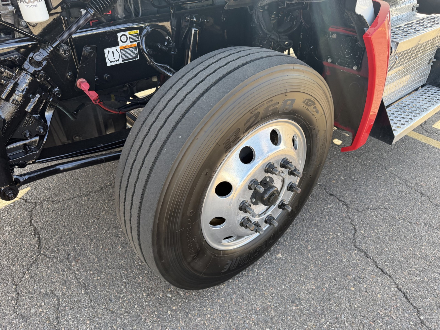 Kenworth T880 front wheel and front step area showing large heavy-duty tire and wheel assembly with hub and lugs; part of the truck's cab and chassis visible.