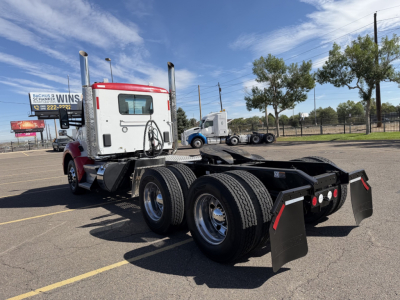 Kenworth T880 tractor chassis with triple rear axles and mud flaps, viewed from rear-left in a parking lot.
