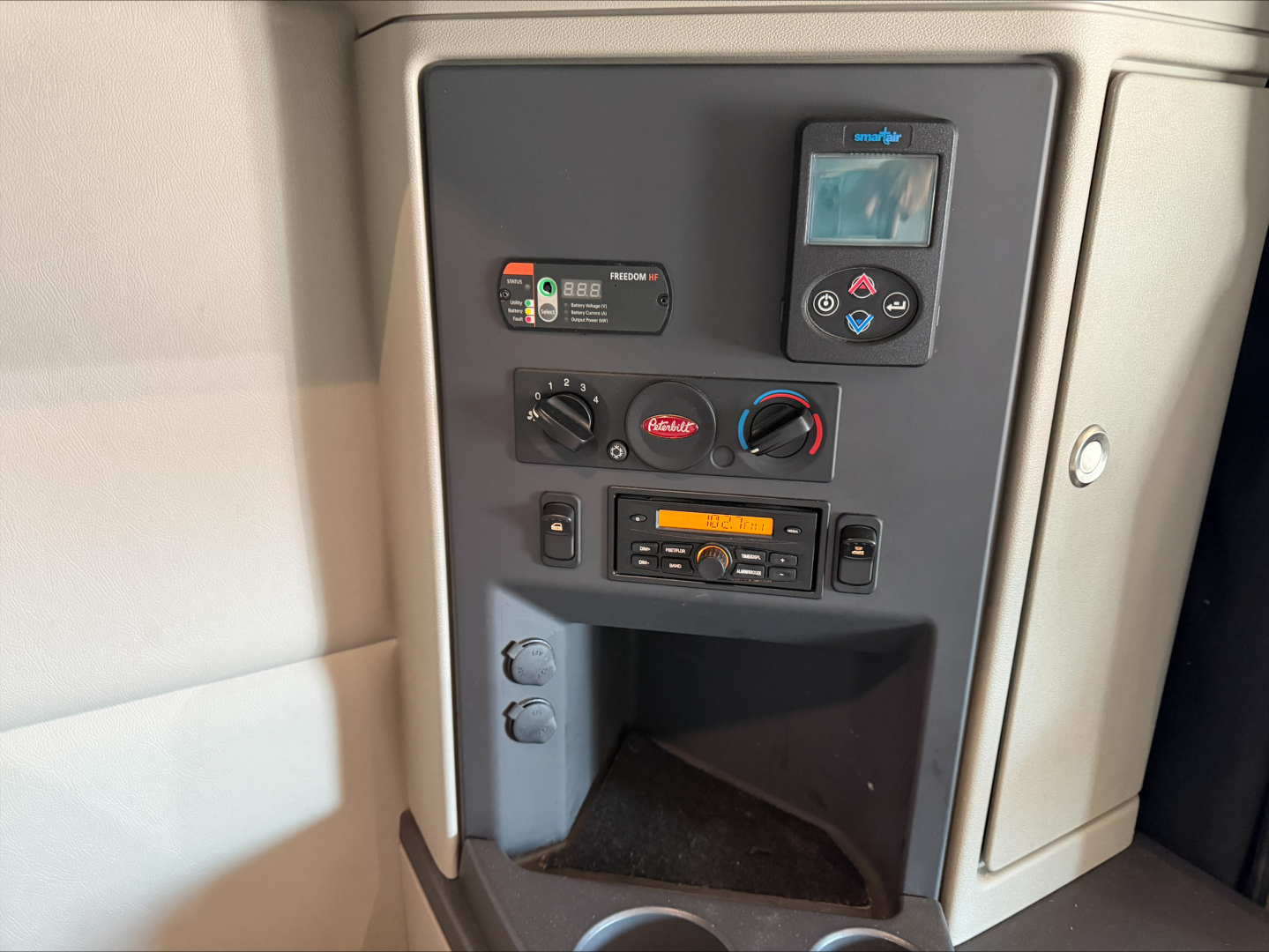 Truck cab interior panel showing a small digital device, a radio, climate controls, and storage compartment. Visible text includes numbers on labels and a small screen.