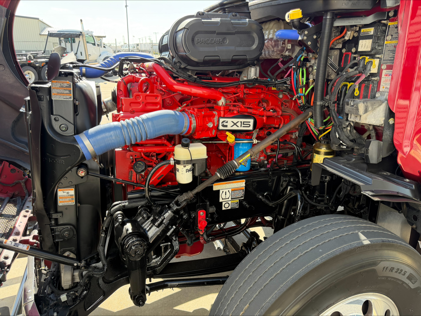 Peterbilt 579 engine bay showing bright red engine with blue intercooler hose, gray air intake cover labeled QMX, yellow dipstick, and wiring harnesses in view.