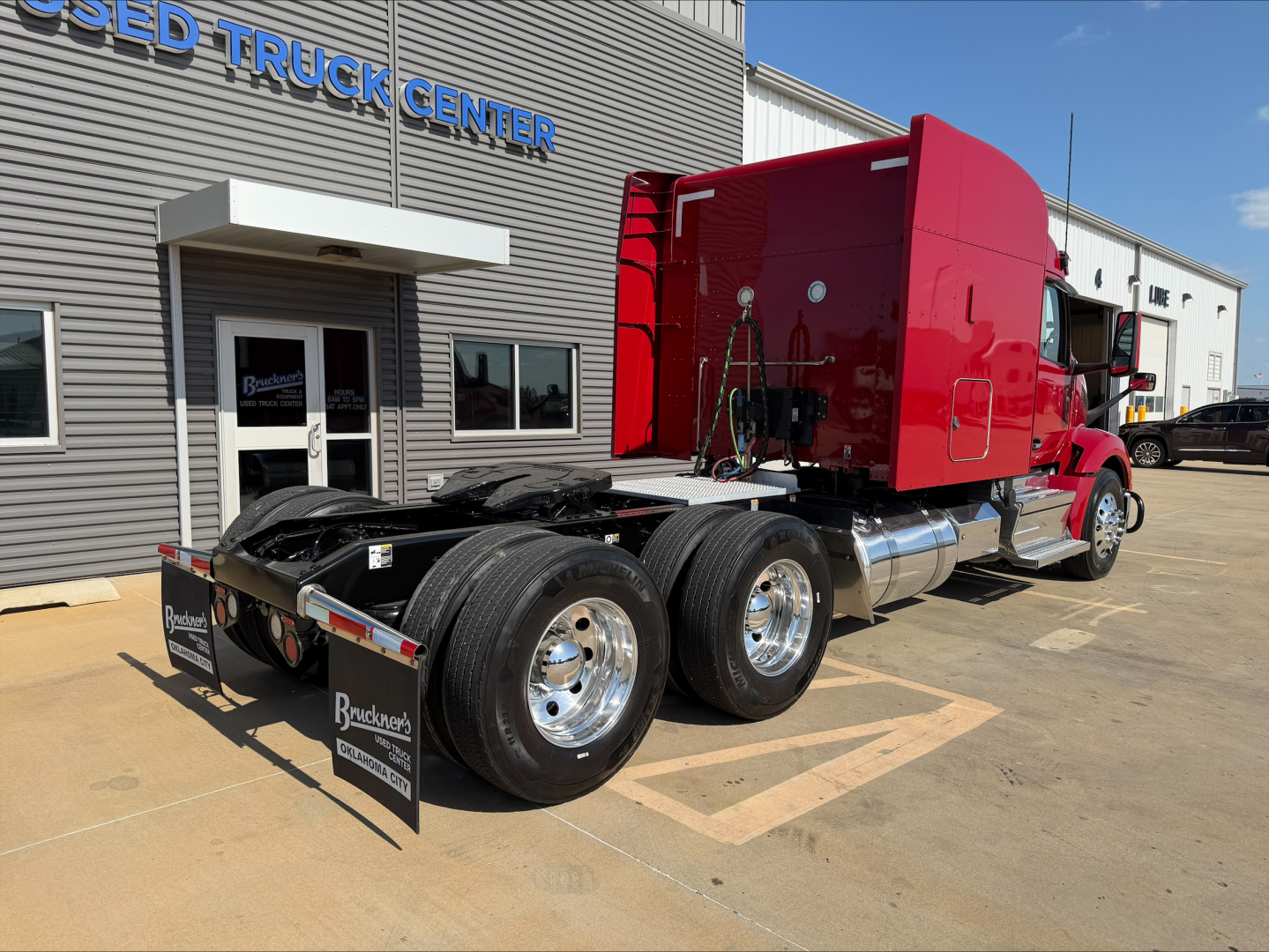 Peterbilt 579 tractor in a truck lot, red cab, dual rear axles without a trailer, chrome fuel tanks, black chassis, stock number B3399P, visible dealership sign.