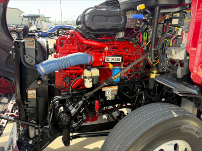 Peterbilt 579 engine bay showing bright red engine with blue intercooler hose, gray air intake cover labeled QMX, yellow dipstick, and wiring harnesses in view.