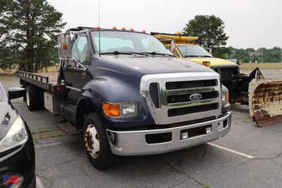 Ford F-650 cab and chassis truck in a parking lot, front view showing dual rear wheels and flatbed, with another vehicle in background.