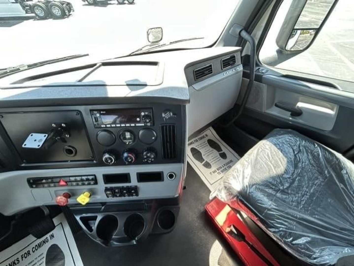 Freightliner Cascadia 126 truck interior dashboard view showing steering wheel, instrument cluster, center console, and driver seat wrapped in protective plastic.
