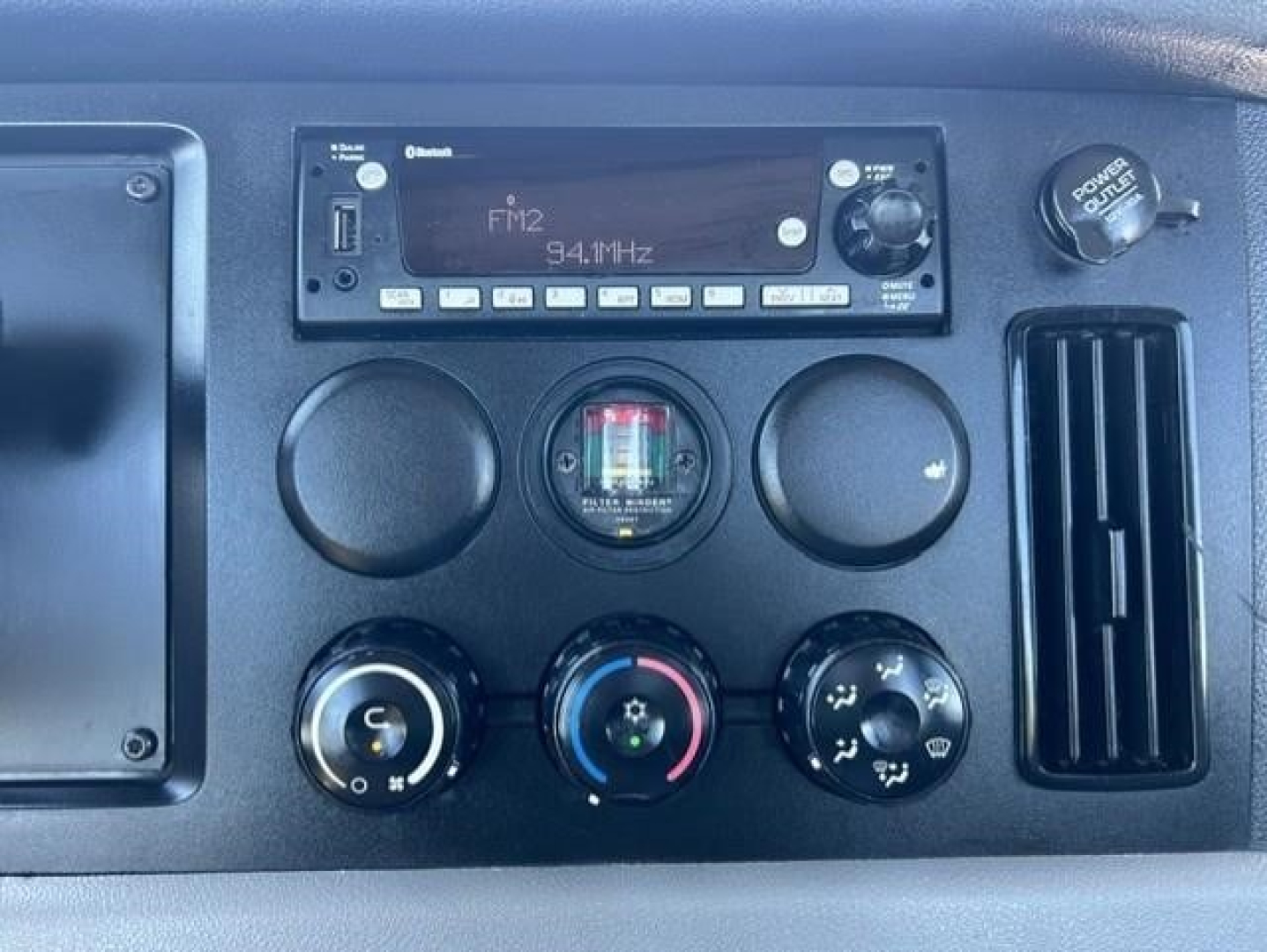 Vehicle dash panel showing FM2 radio with 94.1 MHz and several circular gauges/dials including climate controls and a power outlet, framed by an air vent.