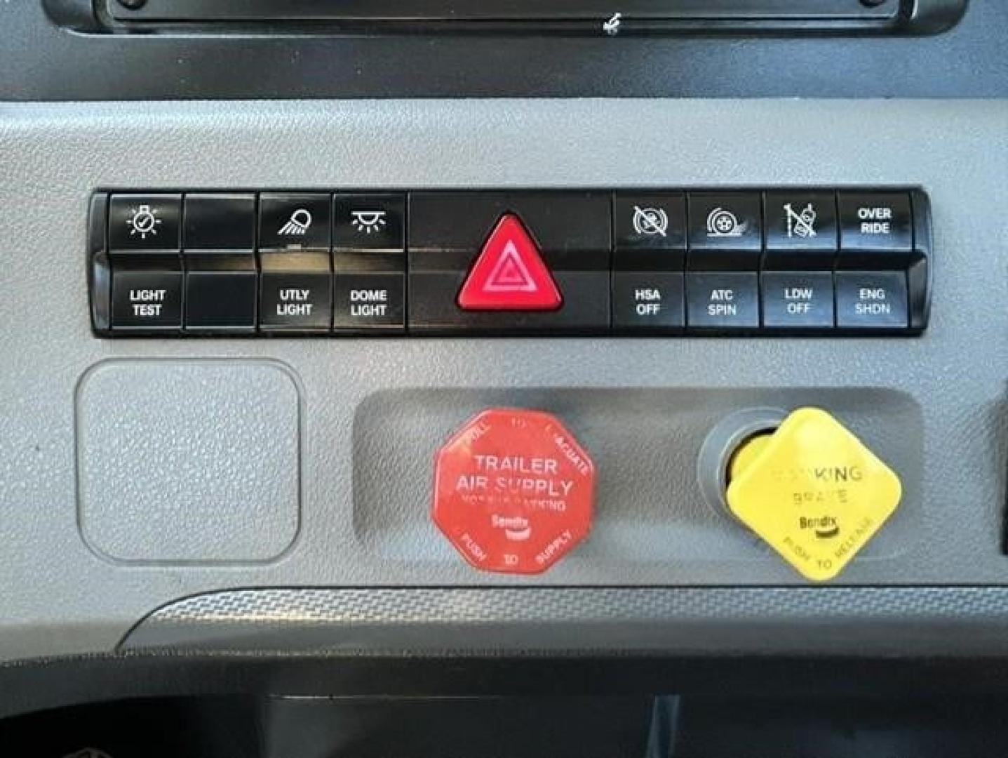 Dashboard controls of a Freightliner Cascadia showing light test, utility light, dome light, hazard switch, and engine off/parking switches with red trailer air supply cap and yellow kingpin cap.