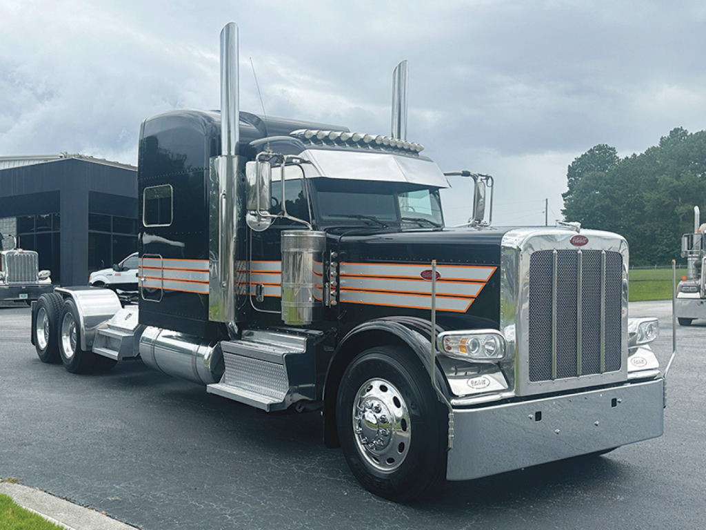 Peterbilt 389 conventional sleeper truck with black paint, chrome grille, and orange pinstripes, front three-quarter view.