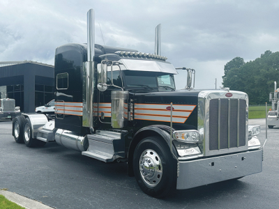 Peterbilt 389 conventional sleeper truck with black paint, chrome grille, and orange pinstripes, front three-quarter view.