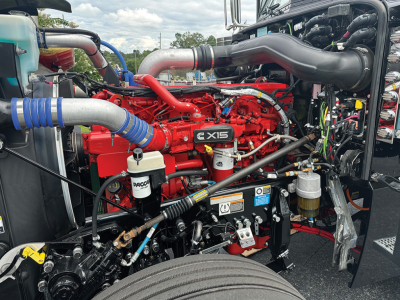 Peterbilt 389 engine bay with red X15 engine, gray intake tubes, blue hoses, and various warning labels visible.