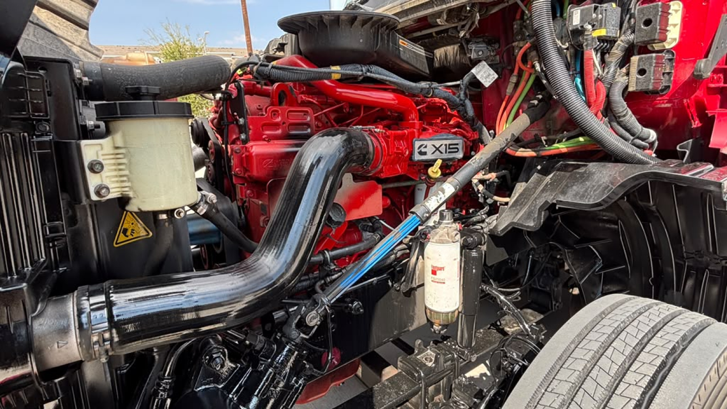 Engine bay showing red Cummins ISX 15 turbo diesel with intake tube and various hoses, white filter housing, and black frame along a sleeper truck.