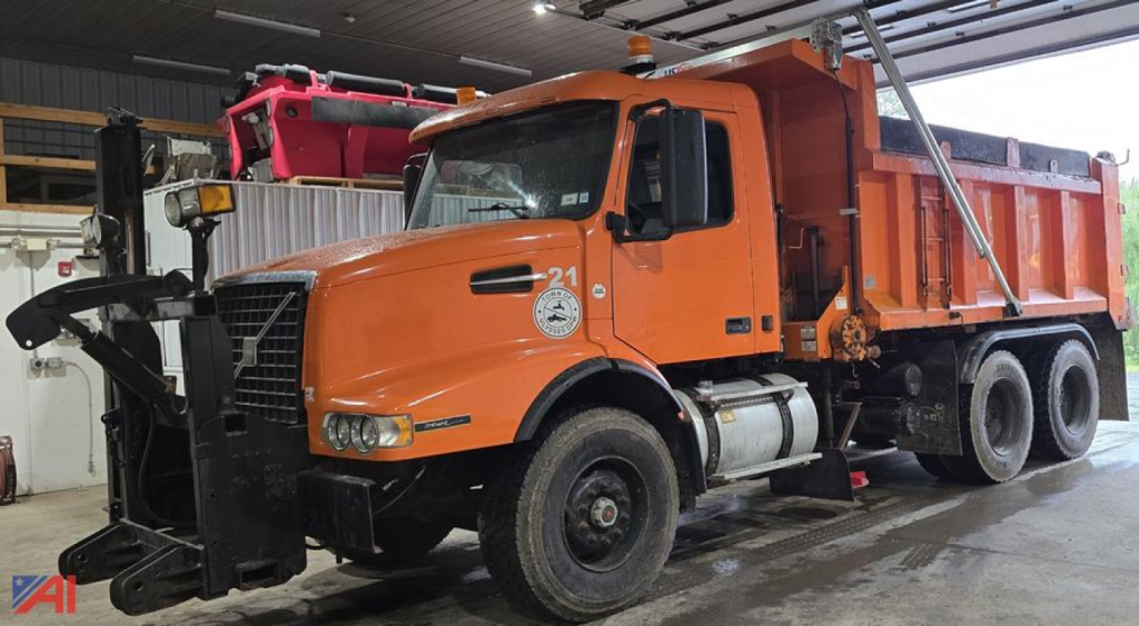 Volvo VHD dump truck in orange with snowplow mounting, showing front grille, cab, and dump bed inside a garage.