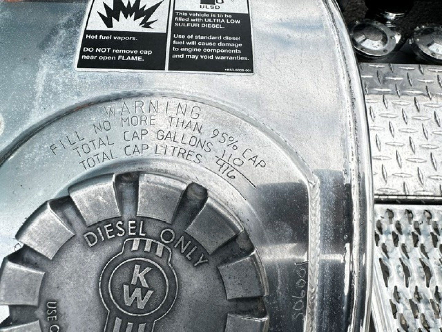 Diesel fuel cap with warning label and warning text: 'FILL NO MORE THAN 95% CAP TOTAL CAP GALLONS TOTAL CAP LITRES 116' and 'DIESEL ONLY' with KW logo. Steps and metal surface visible around pentagonal plate and textured steps.