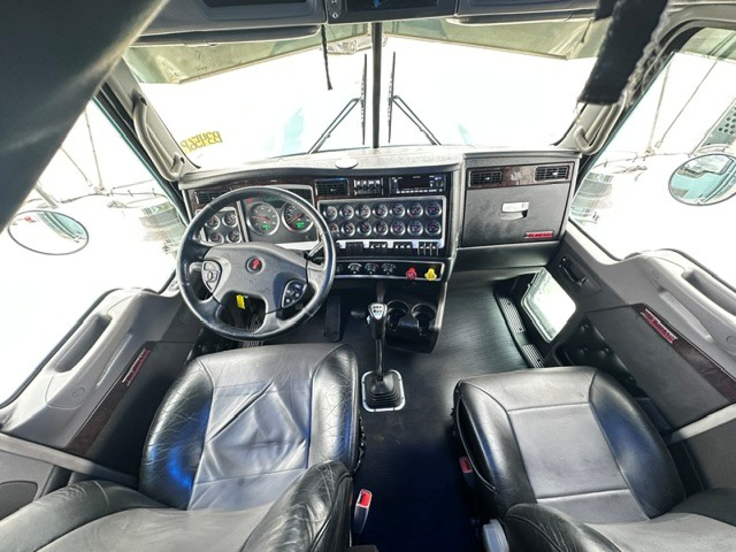 Interior cab view of a Kenworth W900L truck showing steering wheel, dashboard with gauges, gear shifter, and two seats.