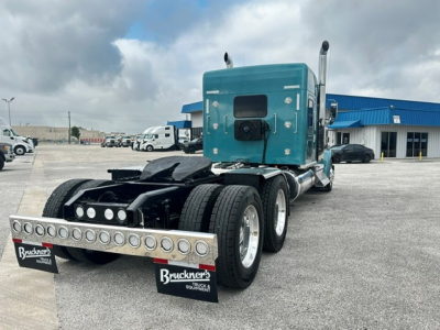 Kenworth W900L tractor rear view showing dual rear axles and flat deck anti-sway bar with round LED light strip; teal cab with sleeper, parking lot, dealership building in background.
