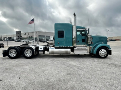 Kenworth W900L conventional sleeper truck side view showing teal cab, chrome exhaust stack, dual rear axles, and gravel lot.