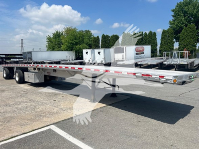 Flatbed trailer shown in an outdoor lot with load deck and side rails. Long trailer with tandem axles and reflective tape along the edges.
