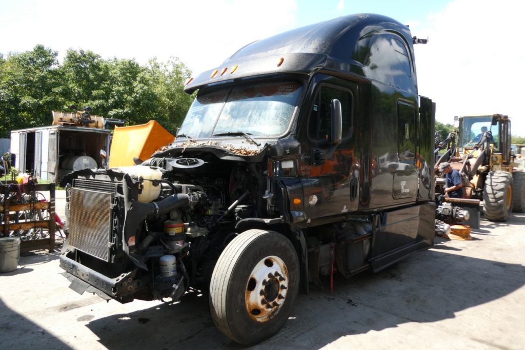 Truck engine bay showing front left view of a damaged 2014 Freightliner Cascadia 125 sleeper cab with exposed engine and components.