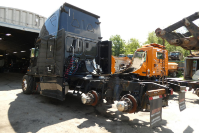 Freightliner Cascadia 125 cab and frame with rear axle components visible, blue cab, orange equipment in background, side view showing exposed chassis.