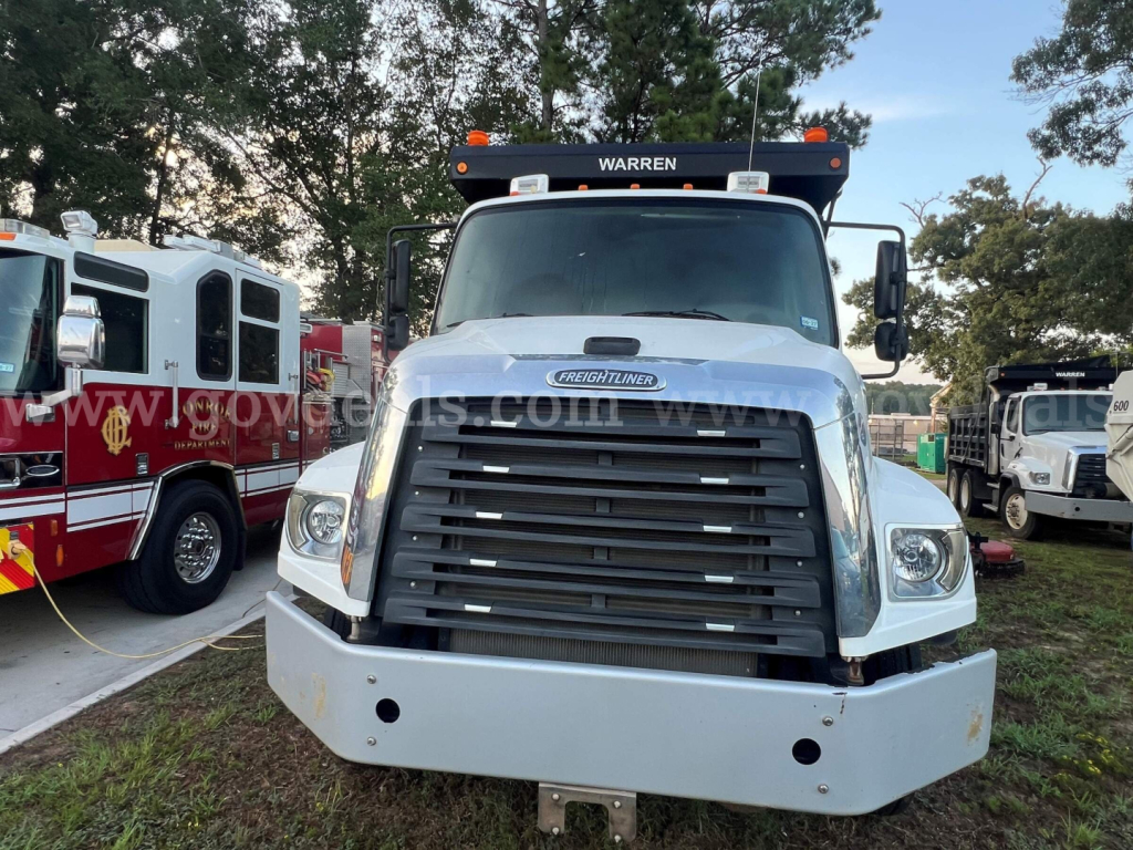 Freightliner 114SD front view showing grille, bumper, headlights, and hood badge. Cab in white, with Warren light bar atop, and adjacent emergency/utility trucks.