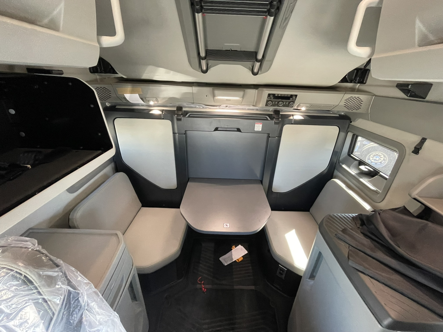 Cab interior of a Volvo truck with a small table, seating area, and overhead storage. Visible details include window, side panels, and a control panel near the top. No legible model or readouts clearly shown.