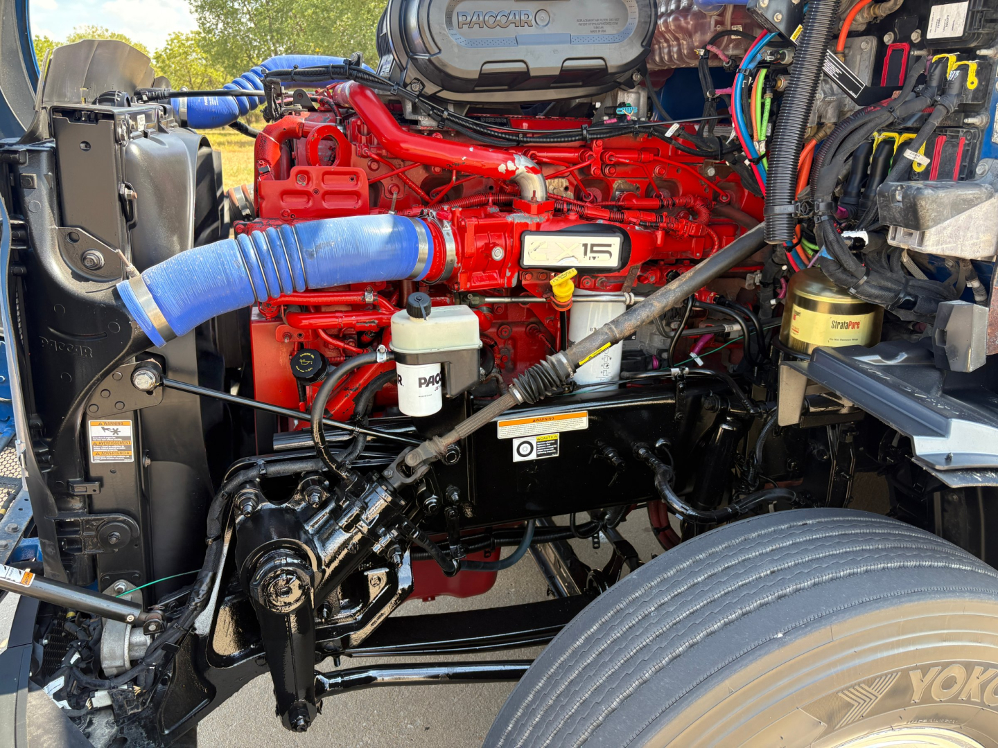 Peterbilt 579 engine bay with red engine block, blue hoses, white brake fluid reservoir, and various electrical harnesses.