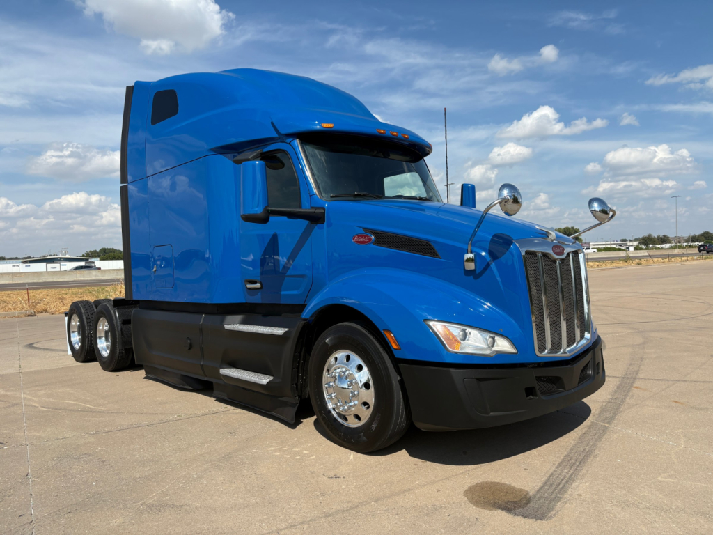 Peterbilt 579 truck with blue cab and sleeper, front view at slight angle, chrome wheels, black bumper, two exhaust stacks, side mirrors, clear sky.