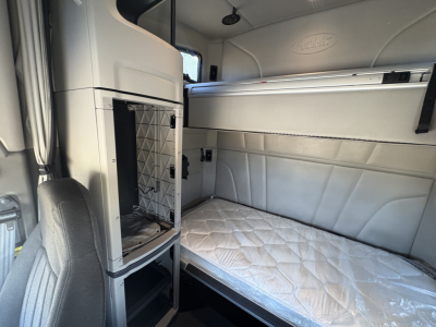 Interior sleeper cab of a Peterbilt truck showing a single bunk bed with quilted mattress and side storage; Peterbilt emblem on panel; overhead cab area and window.