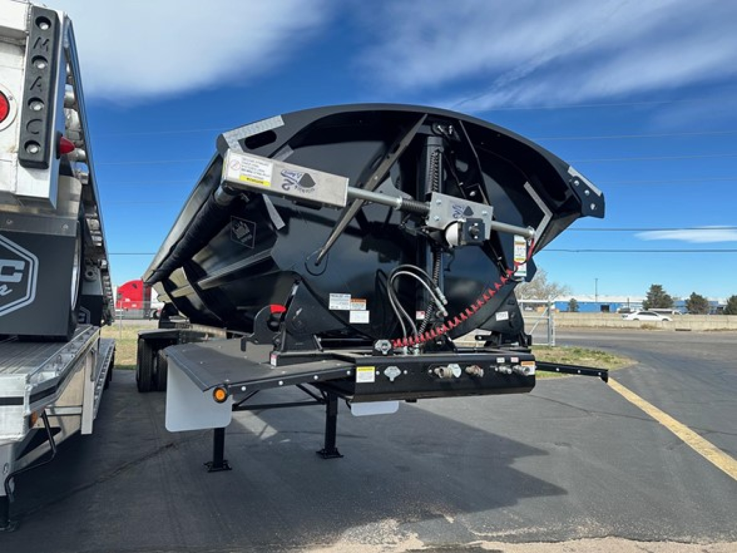 Manac Combo Drop Deck trailer in black with curved rear section, adjacent to another trailer; visible hydraulic hoses, safety chains, and support legs under the deck.