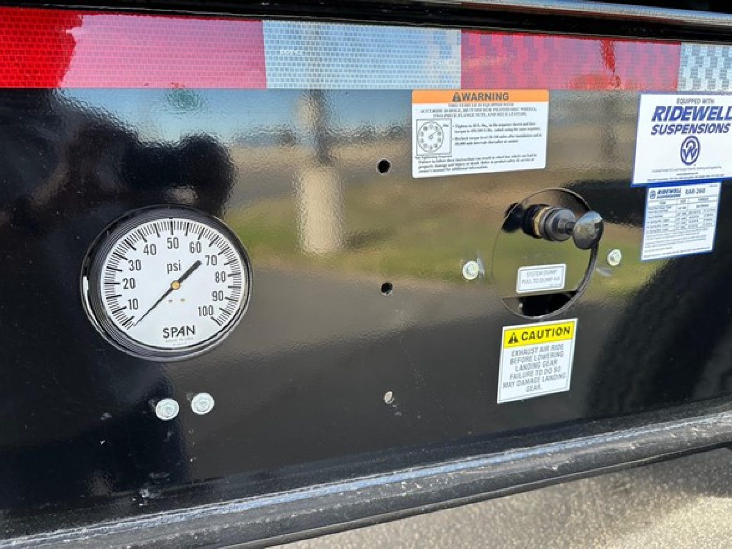 Gauge panel on a Manac trailer showing a pressure gauge with 0-60 psi scale and stickers including WARNING and CAUTION labels, plus a small lever assembly.