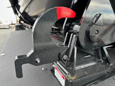 Drop deck trailer hitch mechanism with red latch lever and metal hooks; close-up shows a black painted metal bracket and warning label on the base.