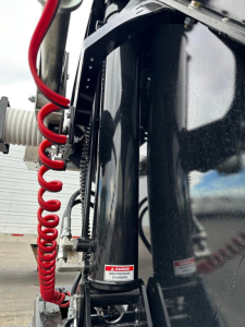 Manac combo drop deck trailer upright frame with red coiled air hose, ladder-like link mechanism, and black glossy side panel; warning label on cylinder area.