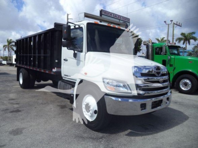 2016 Hino 268A dump truck in a dealership lot; white cab, black dump body, chrome front bumper, large grille, side mirrors, and a green truck in background. Visible signage on top reads 'Truck Sales Center' with palm trees and blue sky.