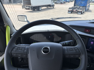 Interior view of a new 2027 Volvo VNL64T860 cab with steering wheel and dashboard; visible Volvo logo on wheel; side mirrors; exterior yard in background.