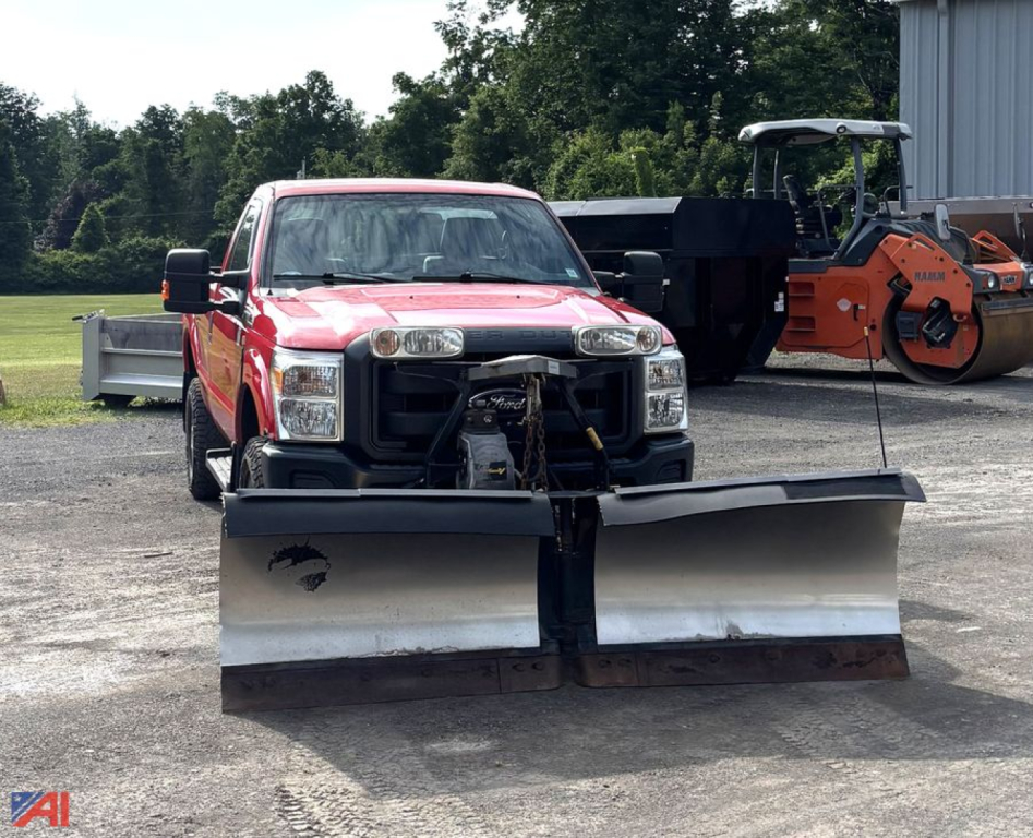 Front view of a red 2016 Ford F-250 Super Duty pickup with a large dual snow plow attached to the front, in an outdoor lot with equipment in the background.