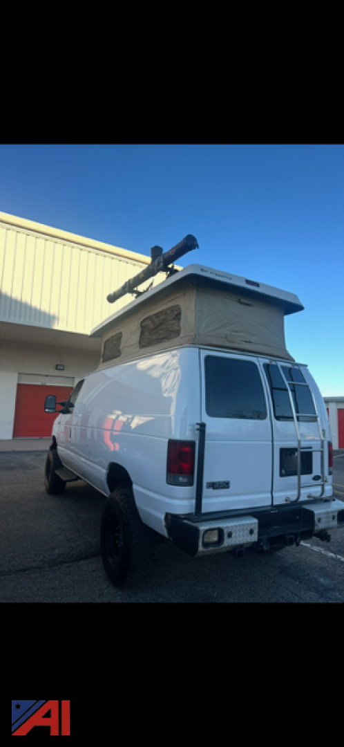 White 2008 Ford E350 cargo van with roof rack and ladder, rear doors, mounted rooftop equipment; visible decals and taillights; parked near a building.
