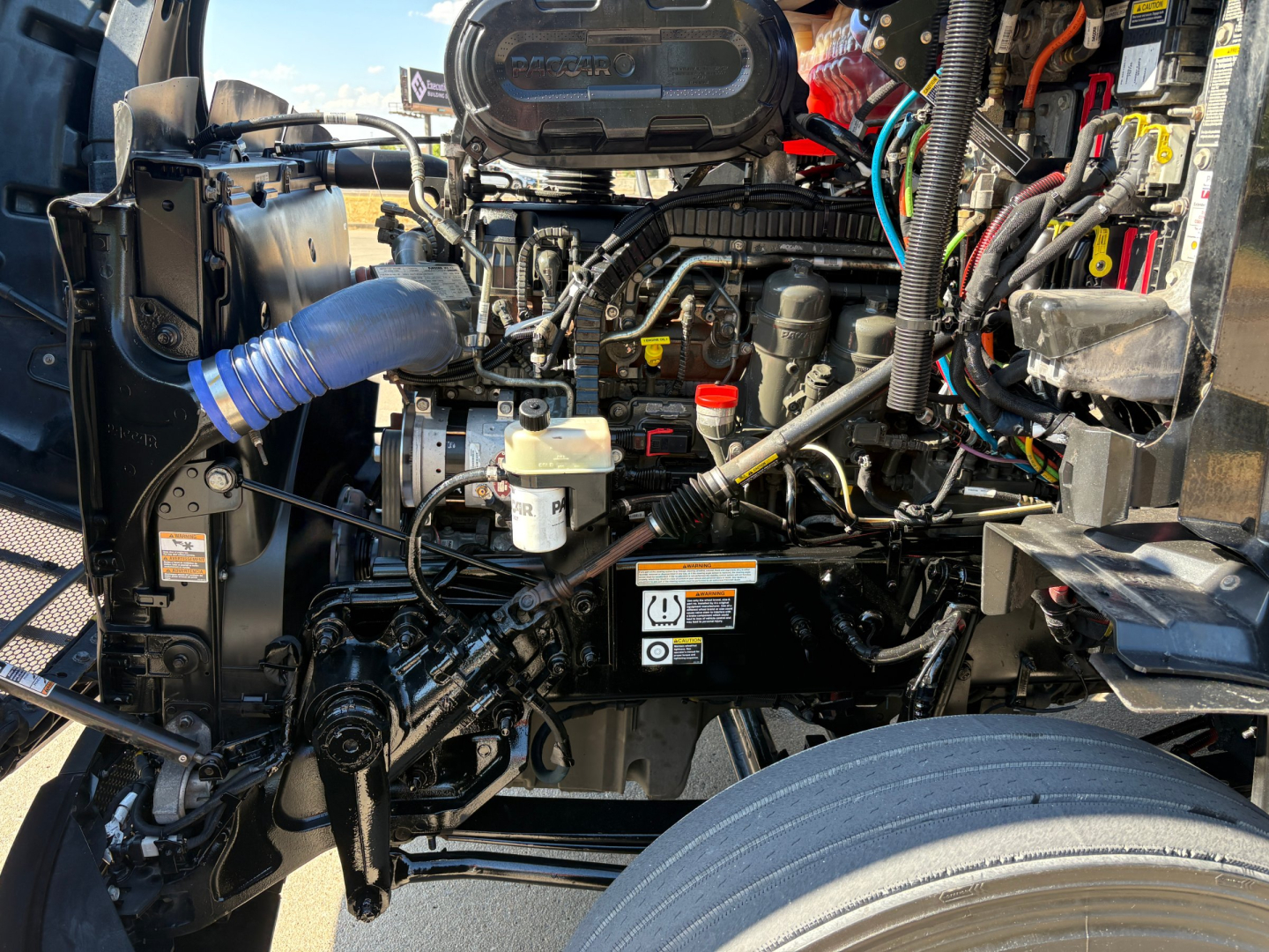 Peterbilt 579 engine bay showing 6- or 8-cylinder diesel engine, various hoses, air intake, battery, and wiring; front wheel visible with fender and suspension components.