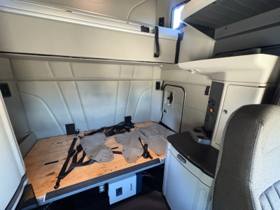 Truck interior side sleeper area of a 2022 Peterbilt 579 showing the cargo floor with a wooden platform and seat/console area in view.