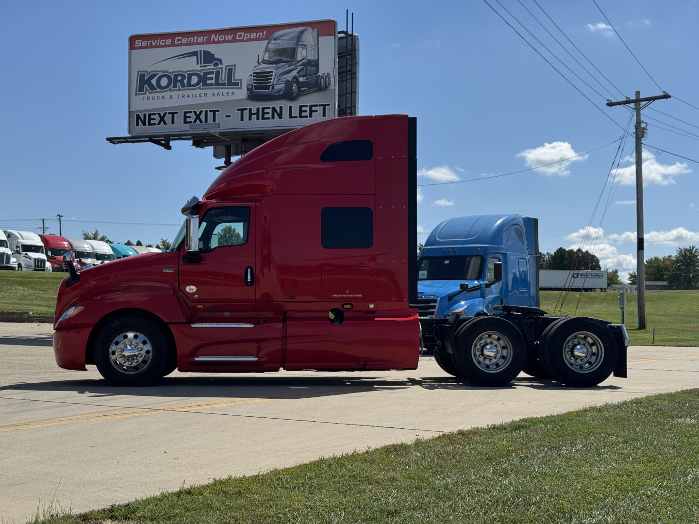 Truck cab and chassis showing red sleeper tractor with blue rear section, parked on lot with billboard in background.