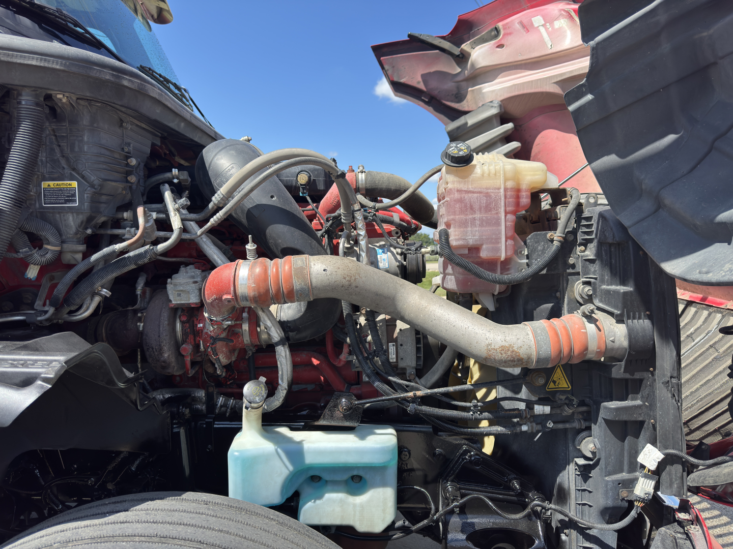 Engine bay of a 2019 International LT625 truck showing coolant reservoir, hoses, and engine components in red and black with surrounding fenders.