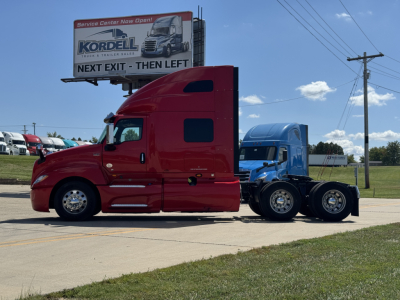 Truck cab and chassis showing red sleeper tractor with blue rear section, parked on lot with billboard in background.
