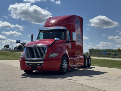 Truck cab with red International LT625 tractor in a parking lot, front-left angle, chrome grille, sleeper cab, sunlight, blue sky with clouds.