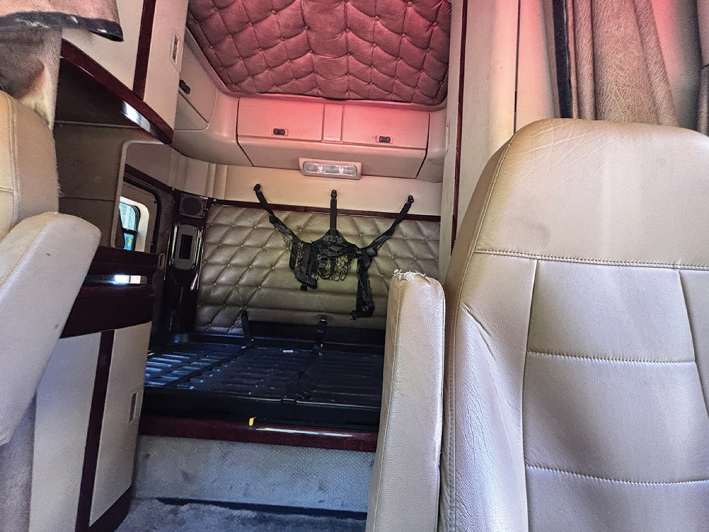 Truck cab interior of a 2013 Freightliner Coronado showing sleeper cab area with beige leather seats and overhead storage; center back area has a black netting and exposed metal framework.
