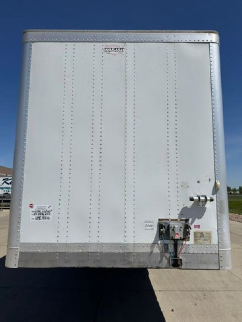 Rear view of a white Wabash trailer: riveted panels, hitch/tongue area with electrical connector, safety chains, and labeling stickers.