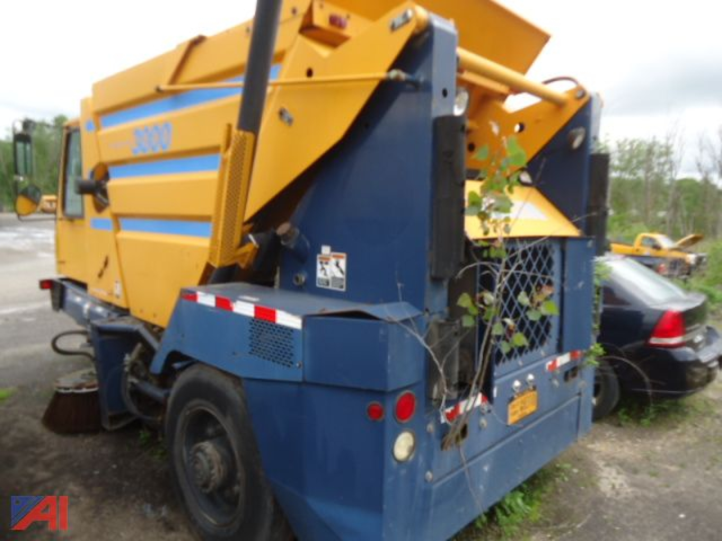 Road sweeper machine showing rear and side with yellow hopper and blue chassis, Johnston 3000 logo visible, debris chute and wheels, rear warning lights.