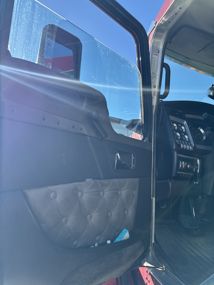 Truck cab door interior of a 2014 Kenworth T660 showing door panel with tufted black upholstery and control area near the dashboard; partial sun glare