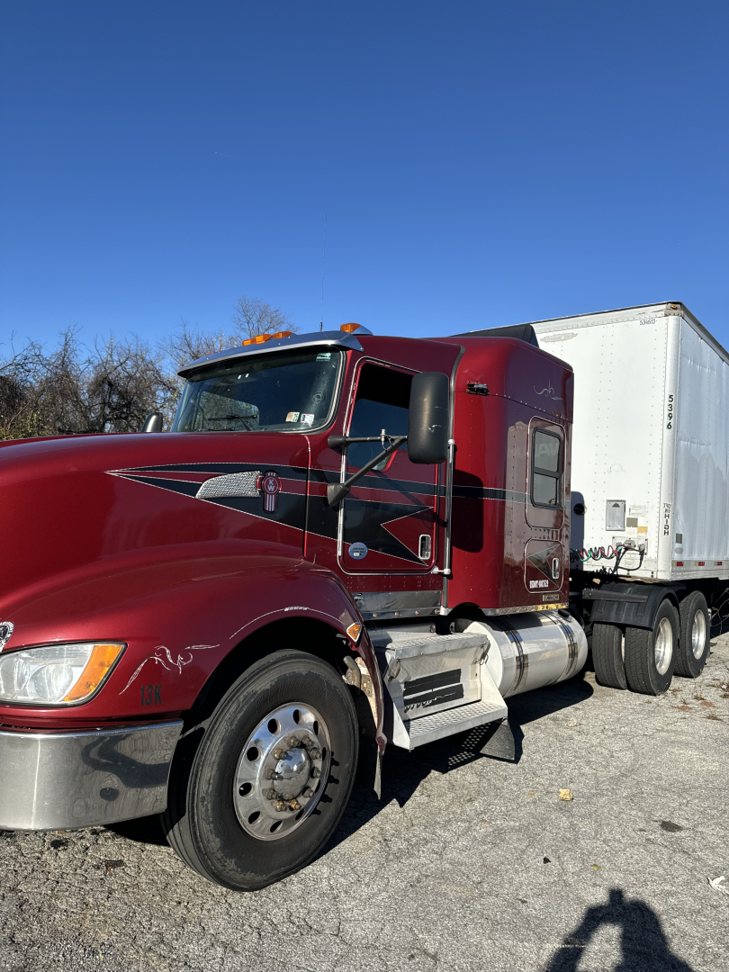 Kenworth T660 tractor-trailer: maroon cab with silver steps, large front wheel, attached white trailer, blue sky in background.