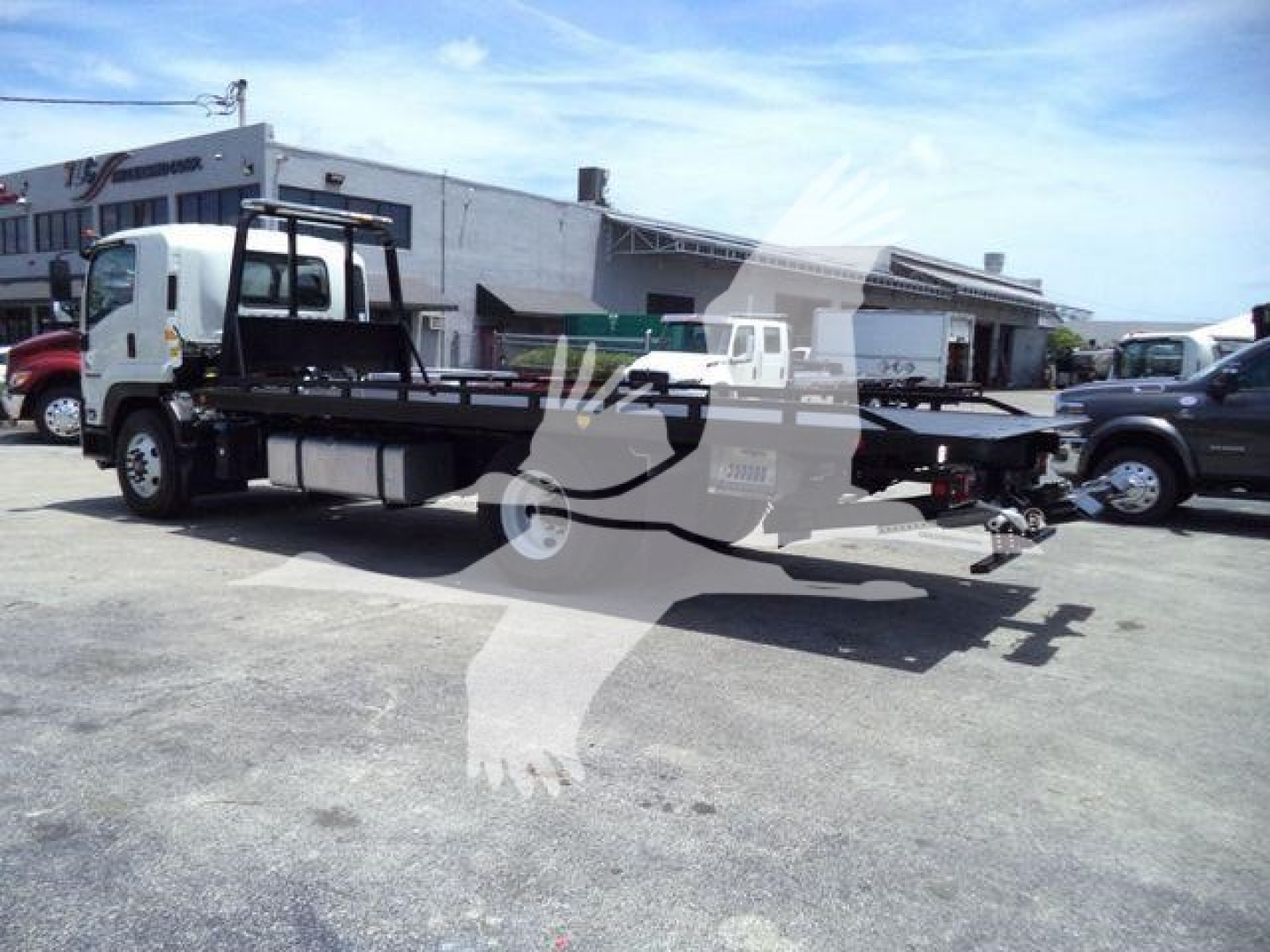 Flatbed tow truck on lot with cab and long flatbed, side view showing chassis and hook lift area.