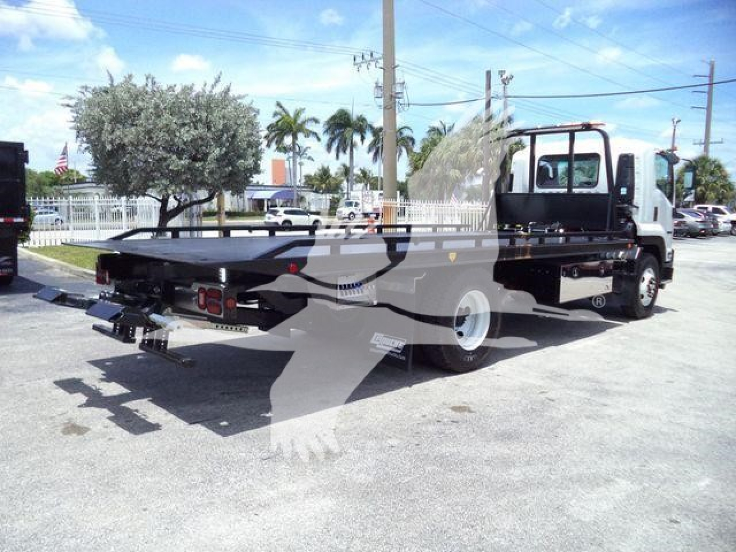Isuzu flatbed tow truck in a dealership lot with rollback equipment and branding on the bed side.